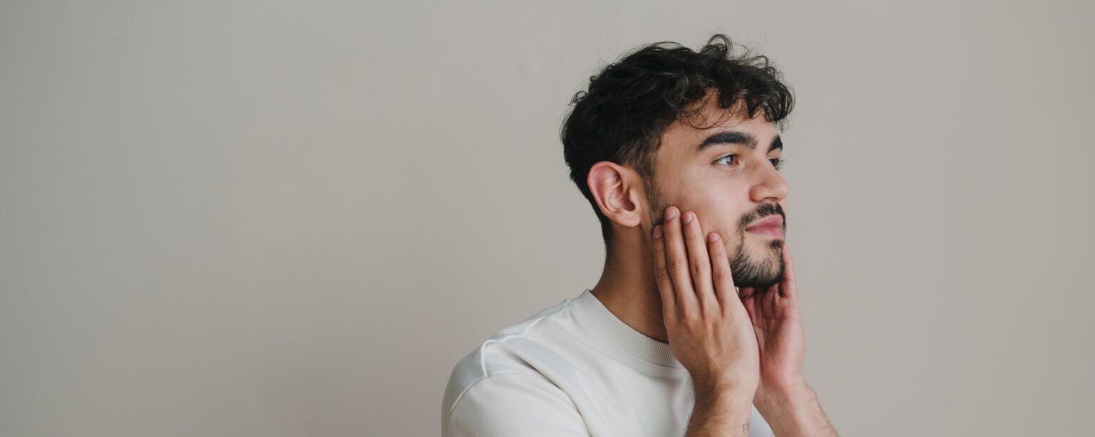 Handsome charming young man isolated over white background, touching his face. Closeup portrait on white background.