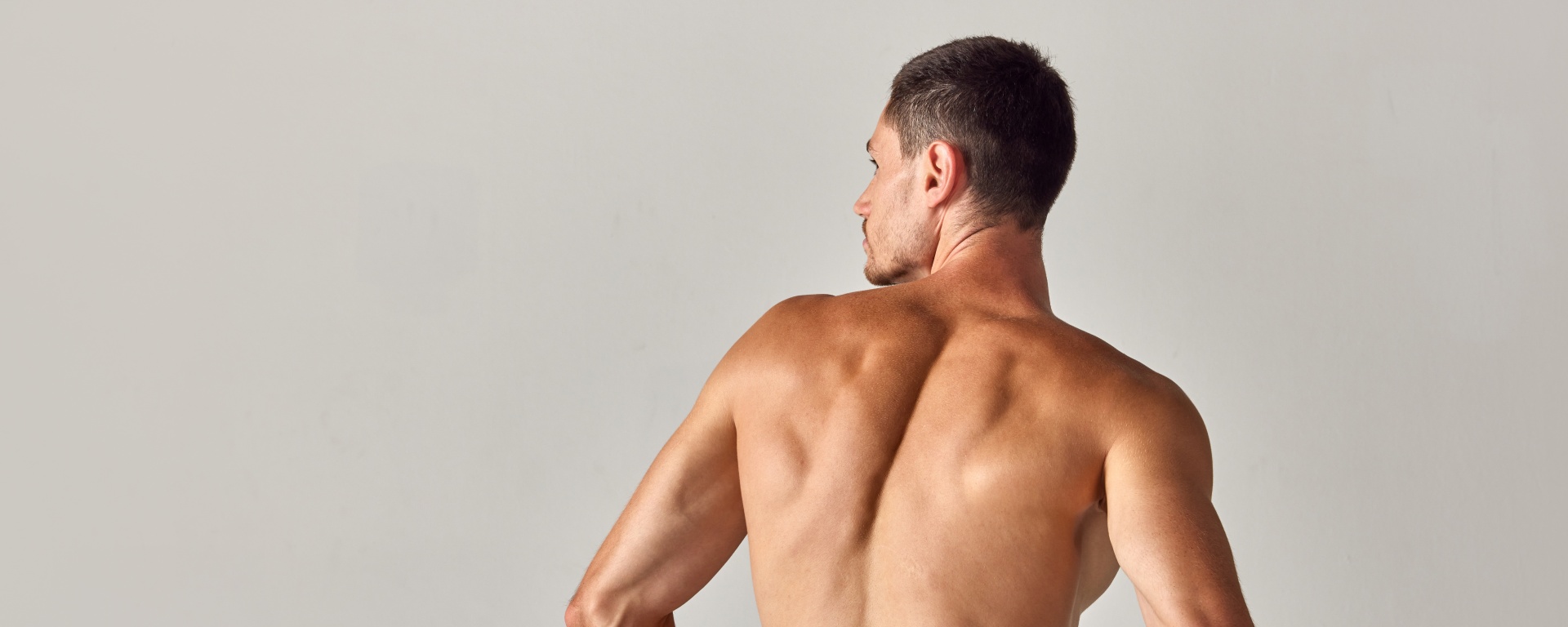 Back view image of muscular young shirtless man with relief, sign back sitting in underwear against grey studio background. Concept of men's health and beauty, body care, fitness, wellness, ad