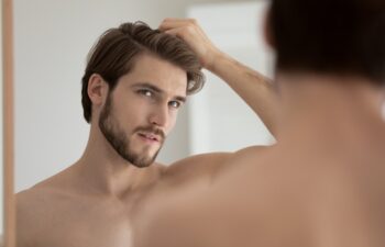 Young shirtless guy looks in mirror touch hair feels concerned due to receding, dry, dull hair. Hair loss common hair problem prevention, alopecia treatment ad, cosmetics products for male concept